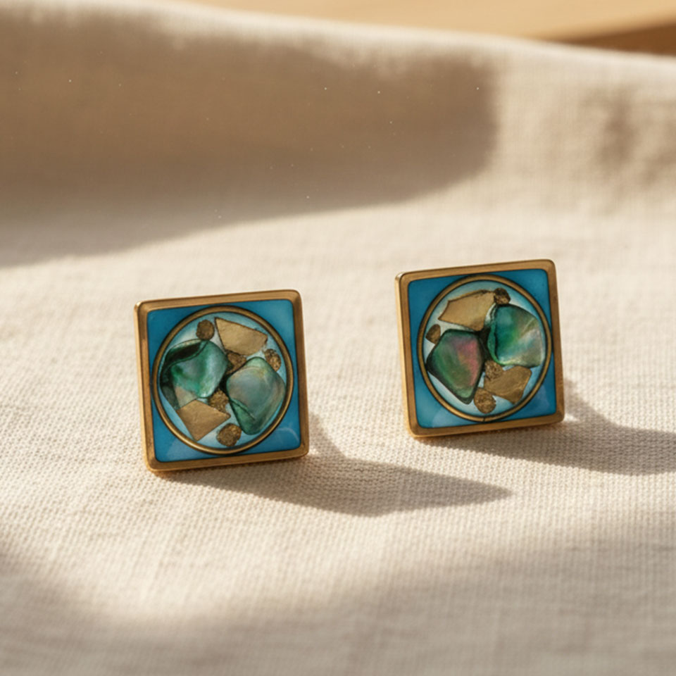 Eleganti e contemporanei, gli orecchini Auralis Cupa uniscono materiali preziosi e design minimale. La composizione in resina racchiude frammenti di madreperla e foglia d’oro, creando riflessi unici che cambiano con la luce. La forma geometrica e compatta li rende perfetti sia per l’uso quotidiano che per occasioni speciali.