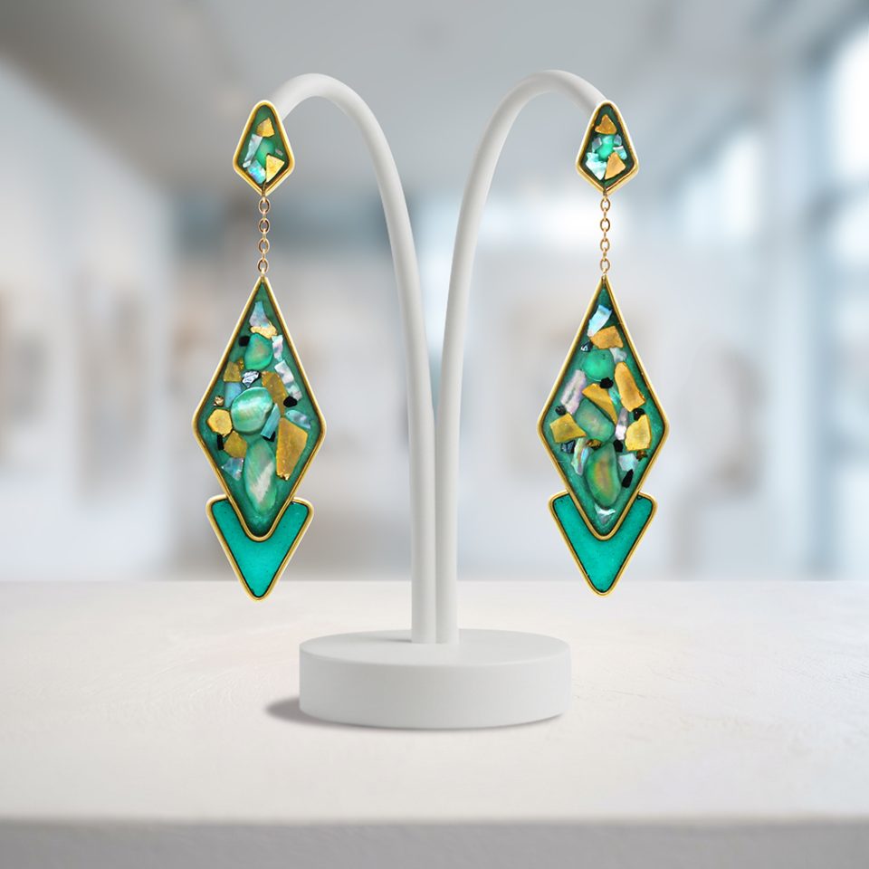 Resina verde smeraldo e turchese con abalone e frammenti di vetro — una palette marina e brillante che cattura la luce ad ogni oscillazione. La catenina dorata che collega i due elementi crea un movimento fluido e naturale, leggero come l'alluminio anodizzato di cui sono fatti. Eleganti e dinamici, impossibili da ignorare.